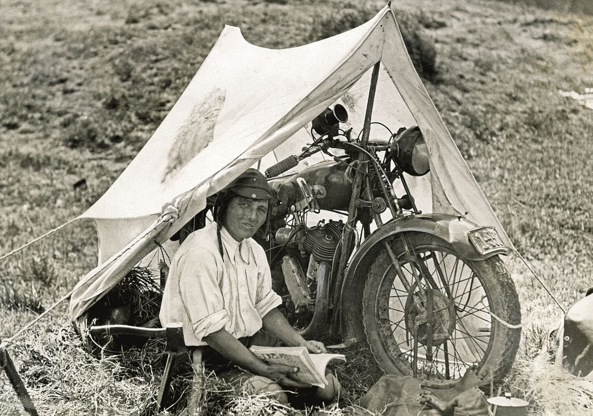 A black and white photograph of Miss Porter looking at the camera as she sits in her tent with her BSA Sloper motorcycle