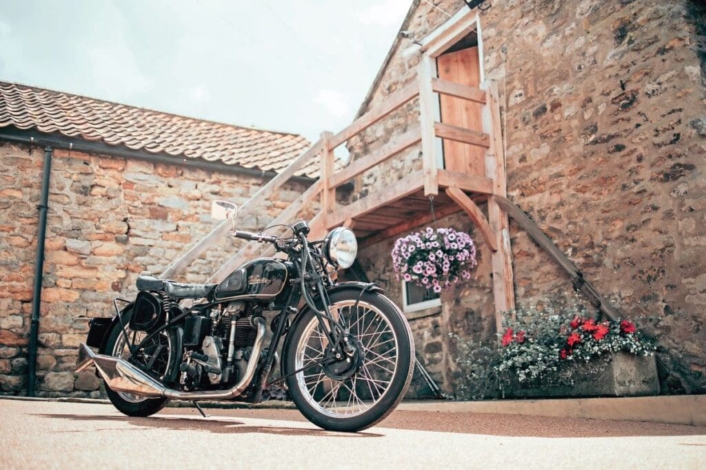 Black Mk.II Velocette KSS in front of brick shed with flowers in the background