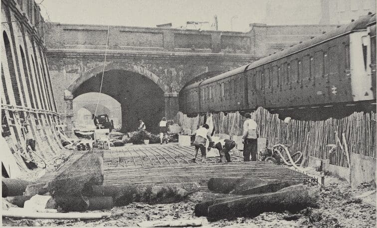Two local tracks—which are to become the fast lines—south of Kentish Town are being relaid on a double concrete slab base at a lower level to give the required clearance under two bridges for overhead wires. Here workmen are laying steel reinforcing material for the lower slab. Passing on the right is a St. Pancras–Bedford diesel multiple-unit train