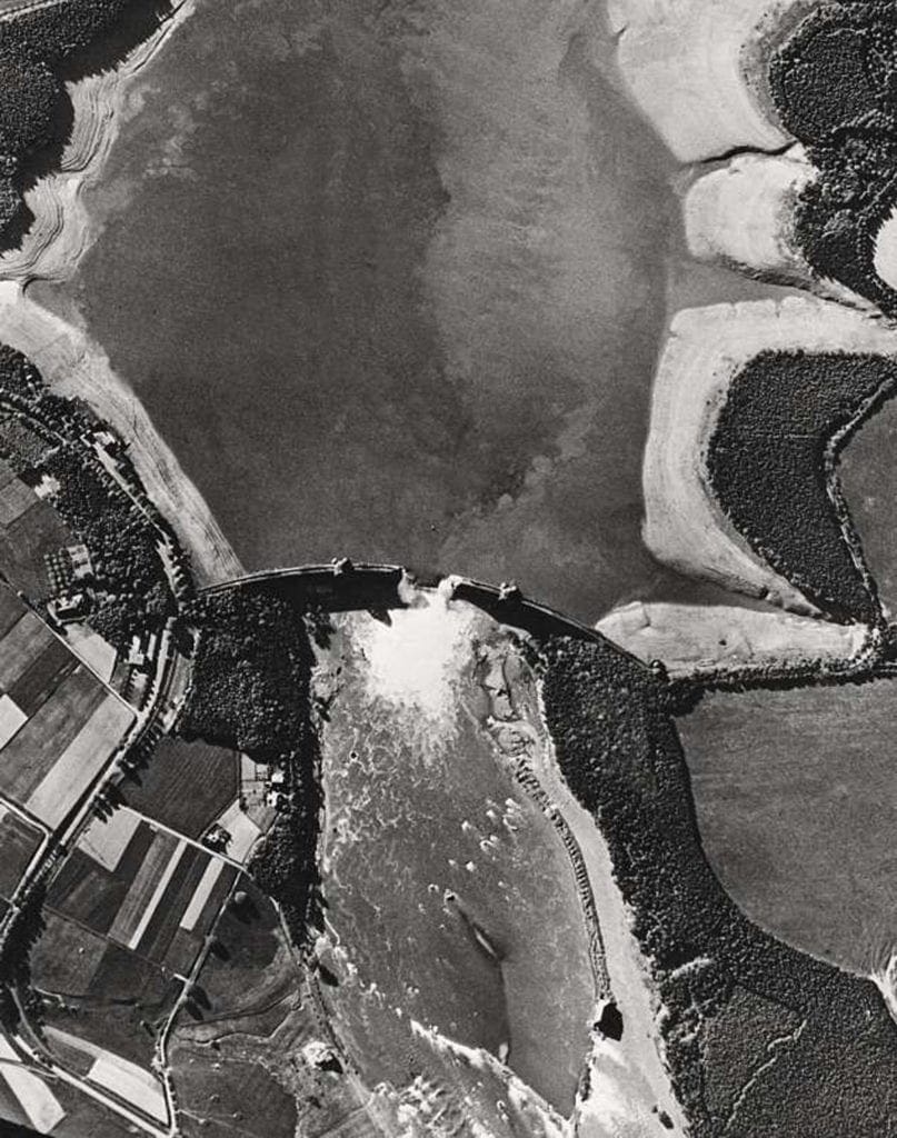 Aerial reconnaissance picture taken on 17 May 1943 clearly showing the breach in the Möhne Dam, water still gushing through and the by then largely drained reservoir. Crown Copyright