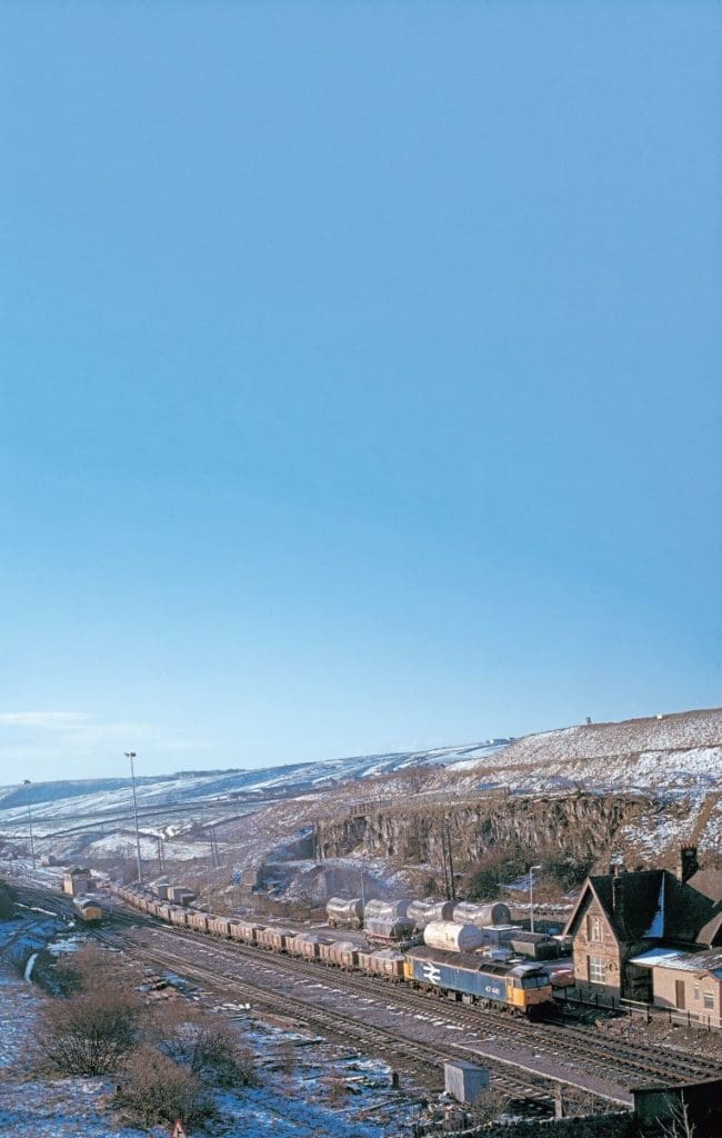 In BR large logo livery, No. 47441 (originally 1557) is seen with a train of two-axle aggregate wagons at Peak Forest on March 2, 1990. Note the two-axle cement wagons in the background and the Class 37 light engine on the left.
