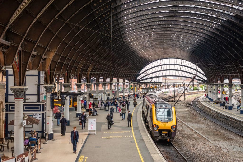 View of a railway station platform with trains waiting to depart and passengers are on the platform. An old curved canopy is above.