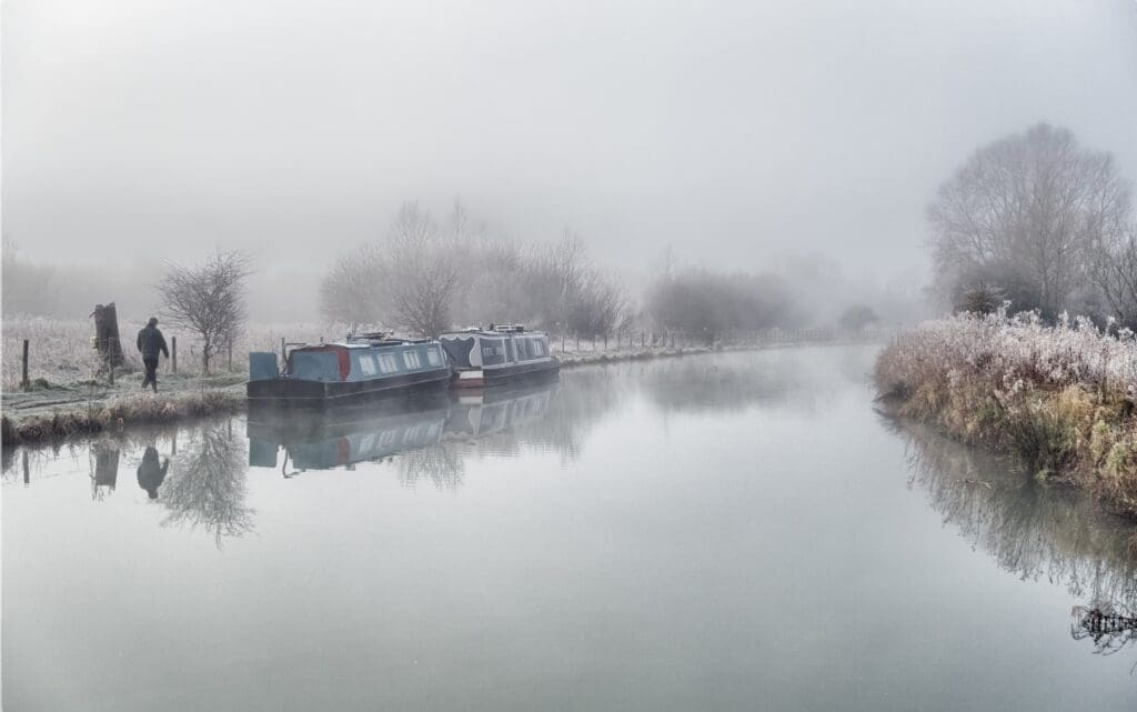 A solitary figure walks their dog along the muddy towpath on the Kennet & Avon Canal, looking west from Hungerford Church, on a cold, misty morning. GILLIE RHODES – CC BY-SA 3.0